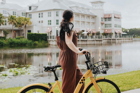 A woman stands beside a yellow cruiser bike along a lakeside path in Celebration, Florida, with pastel waterfront buildings reflected in the calm water behind her.