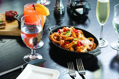 A close-up of a restaurant table setting featuring a dish of fried calamari topped with peppers and herbs, accompanied by drinks including water, a cocktail, and a glass of sparkling wine.