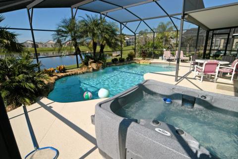 Screened-in private pool and hot tub at a Kissimmee vacation home, featuring tropical landscaping, outdoor dining seating, and a peaceful pond view.