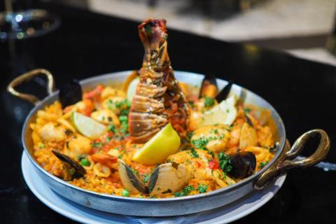 A pan of seafood paella topped with a lobster tail, mussels, shrimp, scallops, lemon wedges, and fresh herbs sits on a white plate on a dark tabletop.