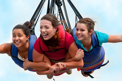 Three women smile and soar together on the SkyCoaster ride at Fun Spot America Kissimmee, suspended high in the air against a bright blue sky.