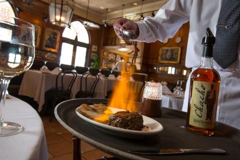 A server in a white uniform prepares a flaming steak dish tableside, with a bottle of bourbon whiskey nearby. The restaurant interior features warm lighting, white tablecloths, and classic décor.