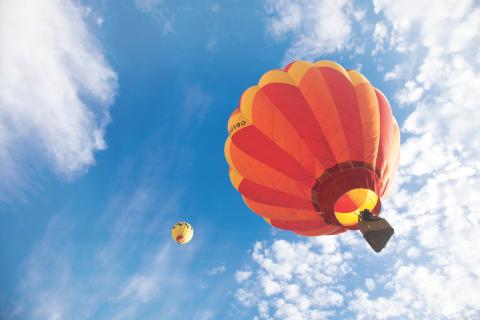 Two hot air balloons—one large and close, one smaller in the distance—float against a bright blue sky with soft, scattered clouds.