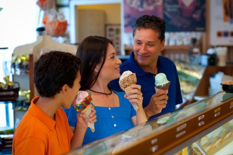 A family inside an ice cream shop enjoying large, colorful ice cream cones, smiling and looking at each other while standing at the display counter.