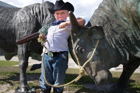 A young boy dressed in western wear poses next to two bulls 