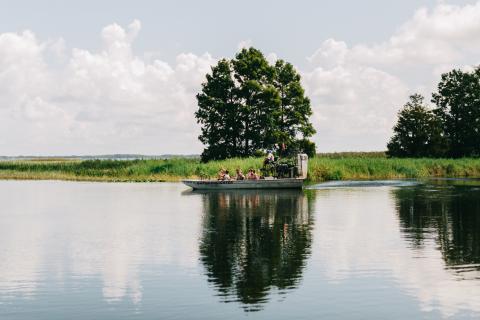 A group of people rides an airboat across a calm lake surrounded by tall grasses and trees under a partly cloudy sky.
