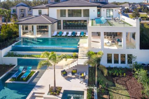 A large modern vacation home with multiple outdoor pools, sun loungers, terraces, and tropical landscaping, viewed from above in a residential resort community.