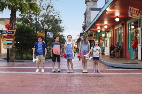A group of six children stand side by side on a brick street at Old Town Kissimmee in the evening, holding balloon animals with string lights and colorful storefronts behind them.