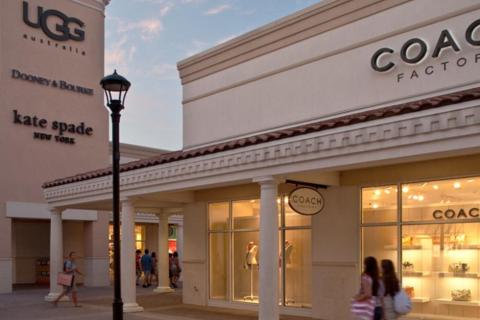 Shoppers walk past storefronts at an outdoor outlet mall featuring brands like Coach, UGG, Dooney & Bourke, and Kate Spade under a soft evening sky.