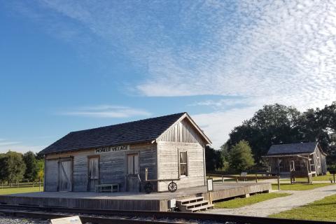 Historic wooden buildings at Pioneer Village sit under a bright blue sky, surrounded by grassy fields, trees, and a gravel walkway.
