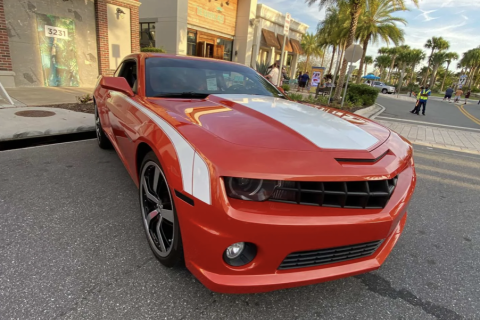 An orange and white muscle car is parked along a street at Promenade at Sunset Walk, with palm trees, shops, and visitors in the background under a bright evening sky.