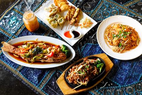 A table covered with a decorative blue patterned cloth holds an assortment of Thai dishes, including a whole fish in sauce with vegetables, shrimp pad Thai, sizzling beef with broccoli, dumplings, spring rolls, chicken skewers, dipping sauces, and a glass of Thai iced tea.
