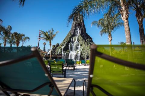 Lounge chairs on a sandy beach face Krakatau Volcano with cascading waterfalls at Universal’s Volcano Bay, framed by palm trees under a clear blue sky.