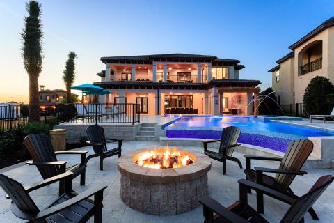 A luxury vacation home backyard featuring a private pool with water features, a fire pit surrounded by chairs, and a spacious patio at sunset.
