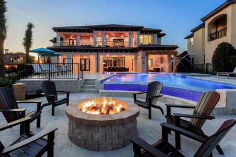 A luxury vacation home backyard featuring a private pool with water features, a fire pit surrounded by chairs, and a spacious patio at sunset.
