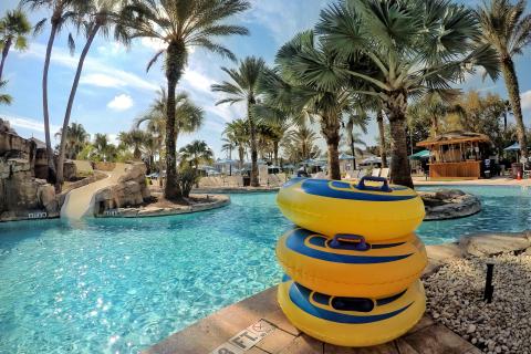 A resort-style pool with clear blue water, a rock waterslide, palm trees, and a stack of yellow inflatable tubes beside the pool deck on a sunny day.