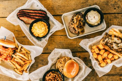 A tabletop spread of barbecue dishes including brisket, pulled pork sandwiches, sides of mac and cheese, baked beans, fries, tater tots, and mashed potatoes from Boggy Bottom BBQ.
