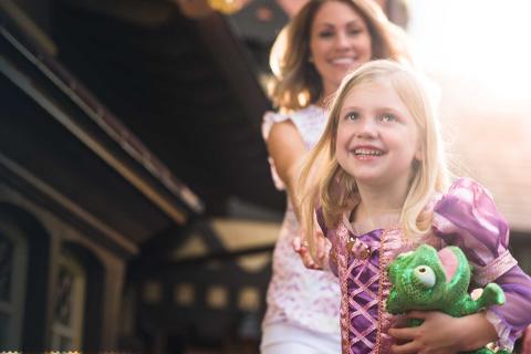 A young girl dressed as a princess smiles while holding a stuffed chameleon toy, with her mother happily walking behind her at a theme park in Kissimmee, Florida.