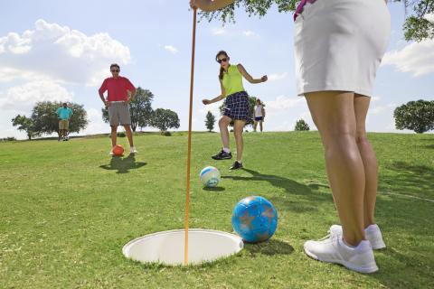 Group of friends playing footgolf on a sunny green, kicking soccer balls toward a large hole on the course.
