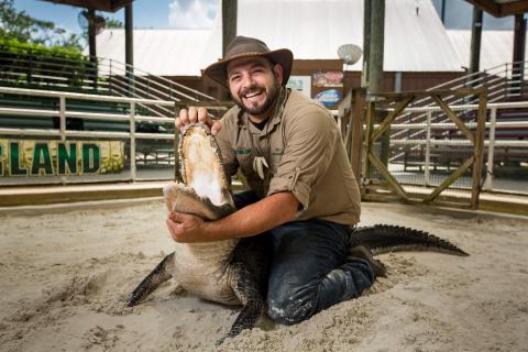 Animal trainer kneeling in a sandy arena at Gatorland in Orlando, Florida, while safely holding an alligator and opening its jaw for an educational demonstration, smiling toward the camera.