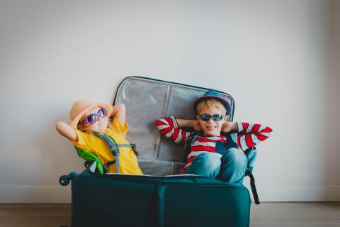 Two young children wearing sunglasses and travel outfits lounge playfully inside an open suitcase, pretending they’re ready for vacation.