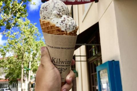 A hand holds a double-scoop waffle cone of cookies-and-cream ice cream in front of a Kilwins storefront on a sunny day.
