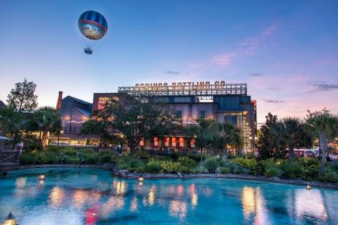 Evening view of Disney Springs with the iconic Aerophile balloon floating above the Springs Bottling Co. building, surrounded by palm trees and water reflecting the sunset sky.