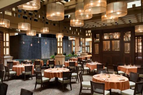 Elegant dining room at Kyng’s Grille featuring warm lighting, round tables with rust-colored linens, and a serene waterfall backdrop.