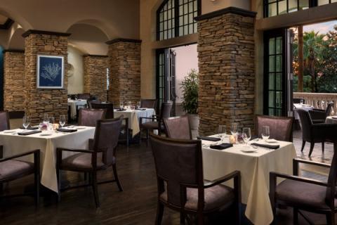 Elegant steakhouse dining room with stone columns, white tablecloths, and large windows opening to a sunset-lit patio.