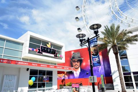 The exterior of Sugar Factory at ICON Park, featuring its colorful signage, outdoor seating area, a large mural of a man in sunglasses and a hat, palm trees, and part of The Wheel visible in the background.