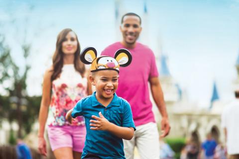 Smiling young boy wearing themed mouse ears runs ahead of his parents at a theme park in Kissimmee, Florida.
