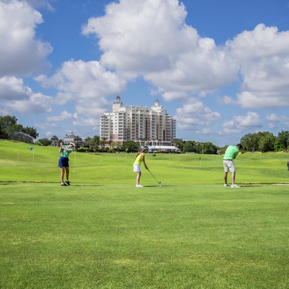 A group of five people practice their swings on a green driving range at Reunion Resort, with a tall resort building and a bright blue sky with scattered clouds in the background.