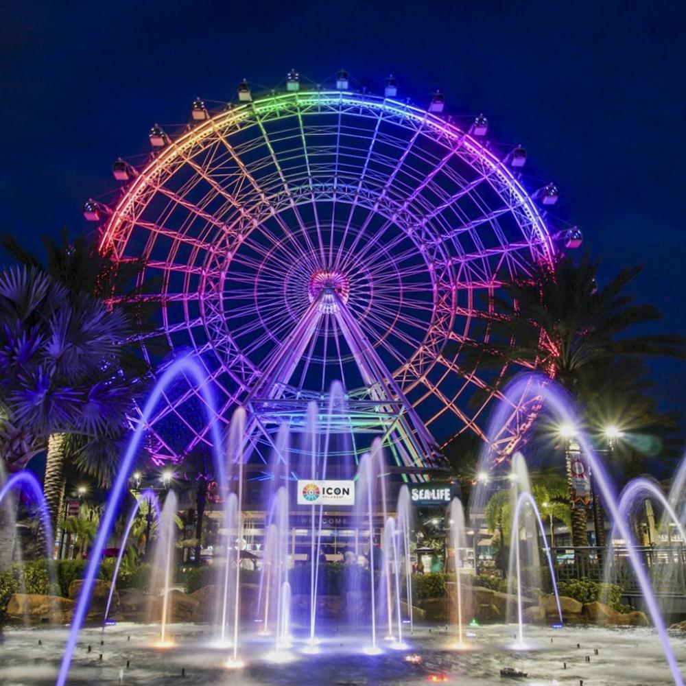 Nighttime view of The Wheel at ICON Park in Orlando, glowing with rainbow lights above a fountain display.