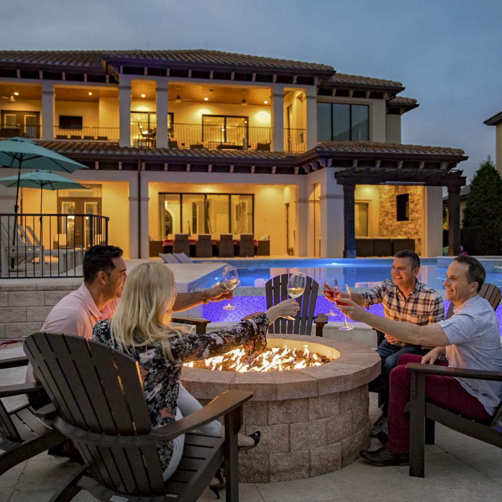Friends gathered around a glowing fire pit at a luxury Kissimmee vacation home, toasting drinks beside the illuminated pool at dusk.