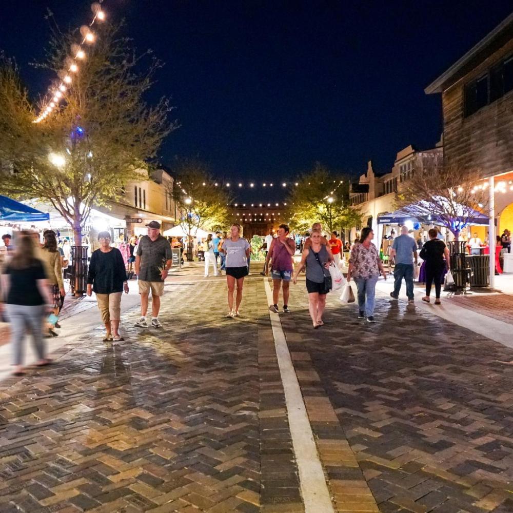 People stroll through a lively nighttime street market in downtown St. Cloud, with vendor tents, warm string lights, and shops glowing along the brick-paved street.