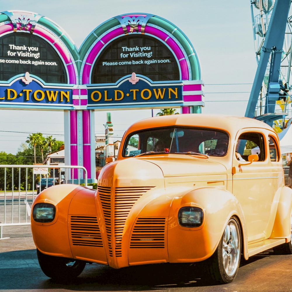 A bright vintage car drives past the colorful Old Town entrance arches in Kissimmee, with the Ferris wheel and visitors in the background.