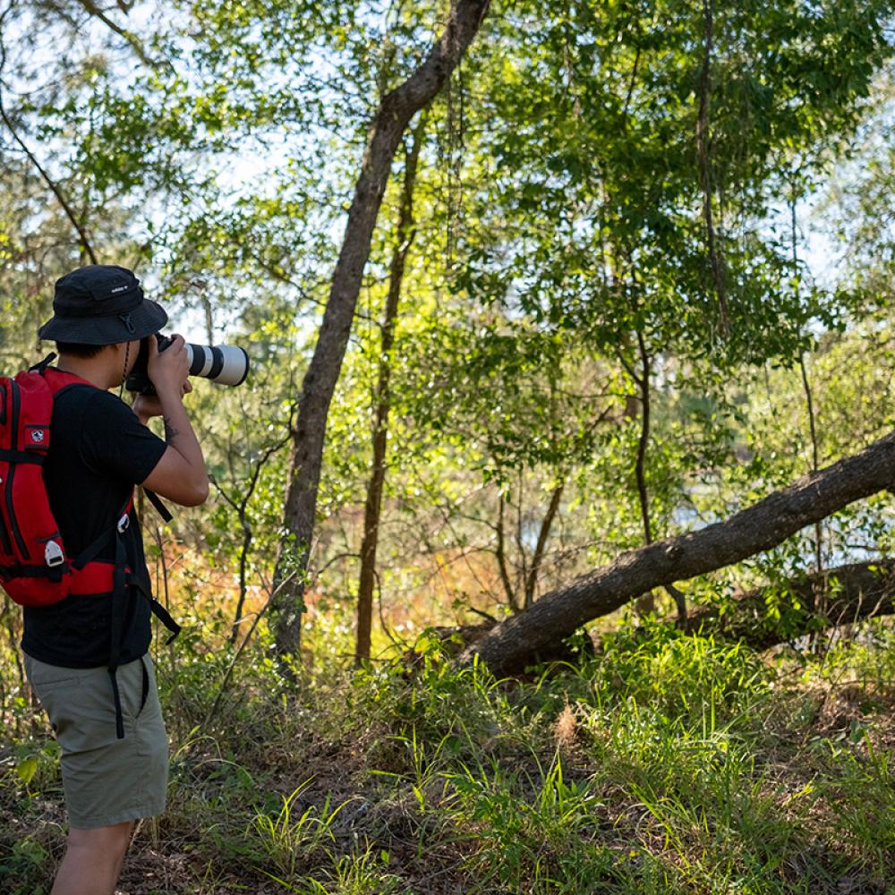 A person wearing a red backpack and bucket hat takes photos with a telephoto lens while exploring a wooded nature area in Kissimmee, Florida.
