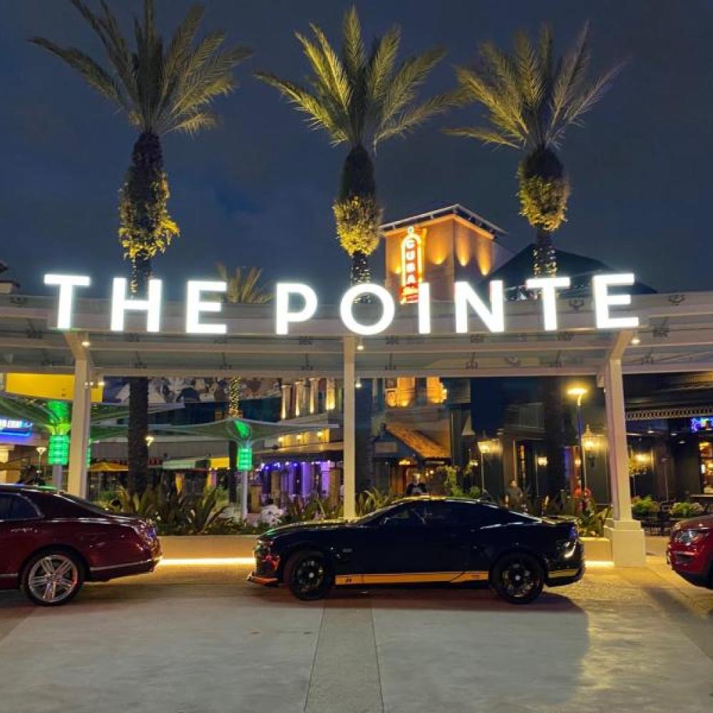 Illuminated “The Pointe” sign glows at night above a valet area lined with parked cars, with palm trees and lively restaurants in the background.