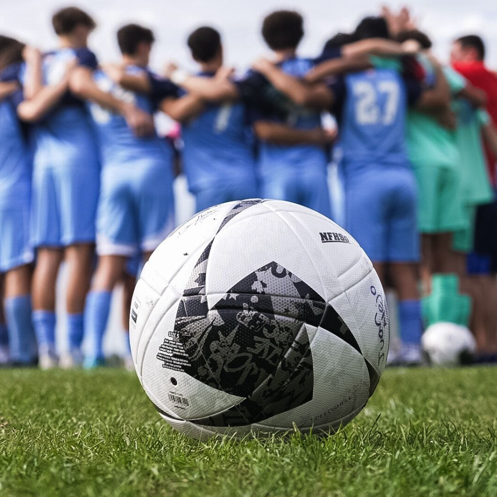 A close-up of a soccer ball resting on the grass with a blurred team huddle of players in blue uniforms in the background.