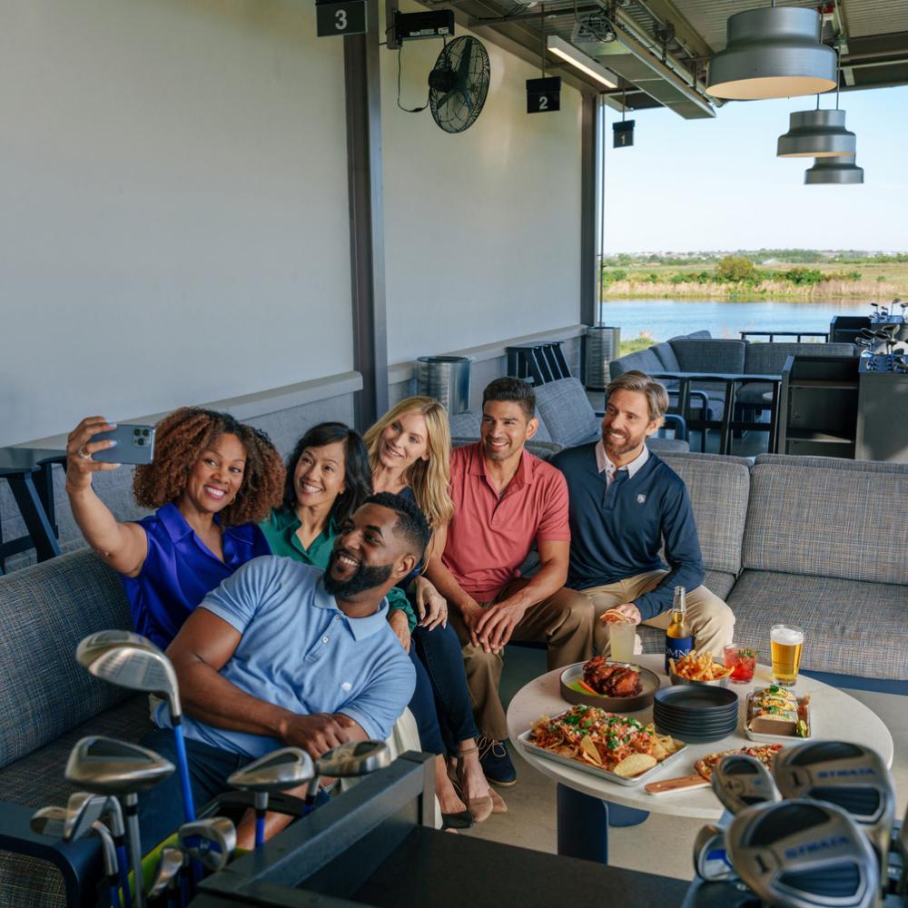 A group of young professionals sit on an outdoor couch closely to take a selfie. In front of them is a variety of small bites and surrounded by golf clubs.