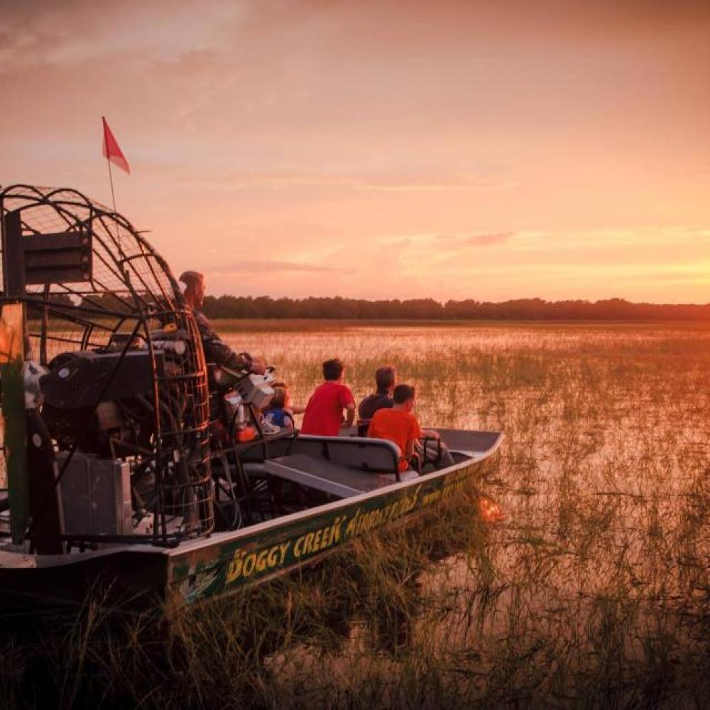 A group of people ride a Boggy Creek airboat through tall marsh grasses at sunset, watching the sun dip below the horizon.