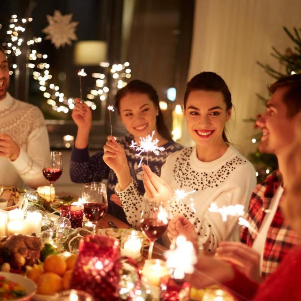Group of friends celebrating the holidays with sparklers and wine around a festive dinner table decorated with candles and Christmas lights.