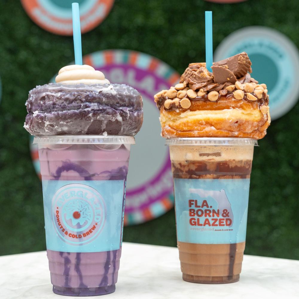 Two drinks in plastic cups are on a white table with a greenery background. The ube drink on the left is light purple with darker purple swirls and is topped with a glazed ube doughtnut and white icing. The peanut butter drink on the right is light brown with chocolate swirls and is topped with a glazed doughnut covered with crumbled peanut butter cups and a straw.