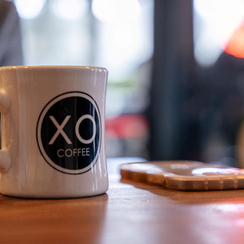A white ceramic coffee mug with a black circle and the words, XO Coffee in white letters in the center, on a wooden tabletop. In the background, there is a faint outline of a neon sign.
