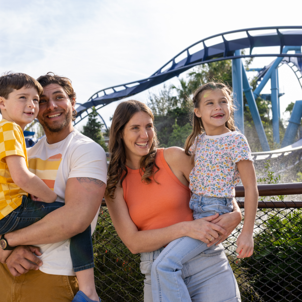 A family of four smiles and poses together in front of a looping roller coaster at a theme park, with parents holding their two young children.