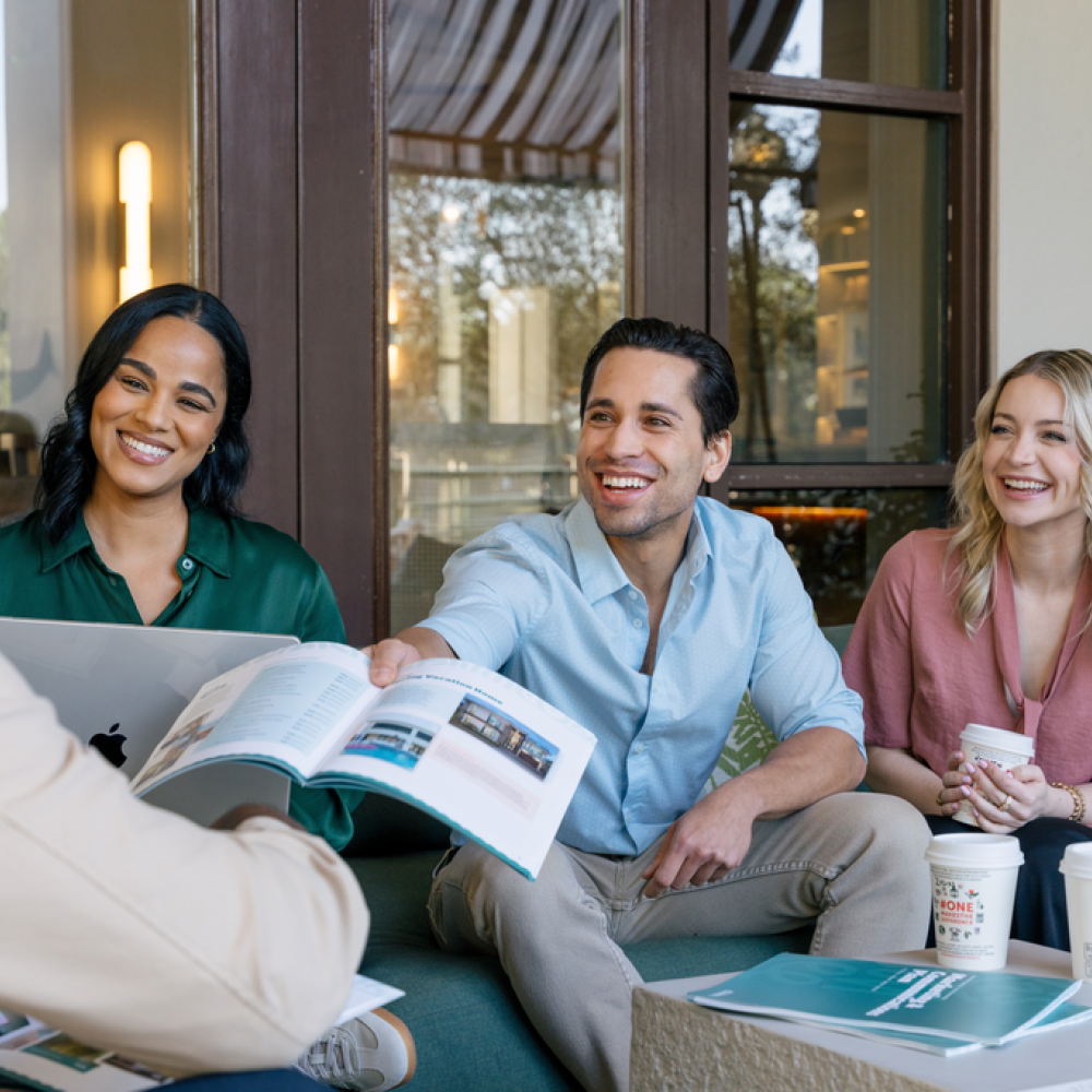 A small group of colleagues sits together outdoors, smiling and reviewing printed materials during an informal meeting.