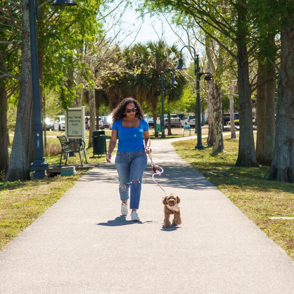 A woman walking her small brown dog on a sunny day at Dan Tarrell Memorial Point in St. Cloud, Florida, with trees lining the paved path.