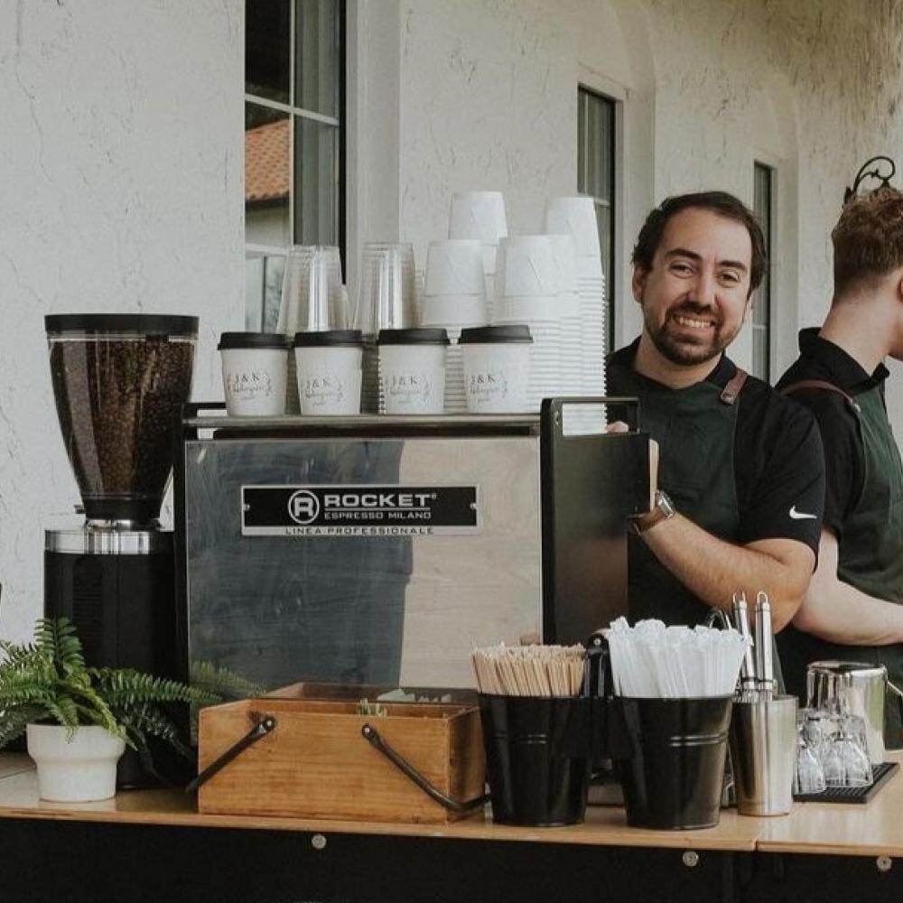Two baristas at Piccolo Coffee Co.’s mobile espresso bar, smiling and preparing drinks during an outdoor event in Central Florida, with a full setup including syrups, coffee grinder, and custom cups.