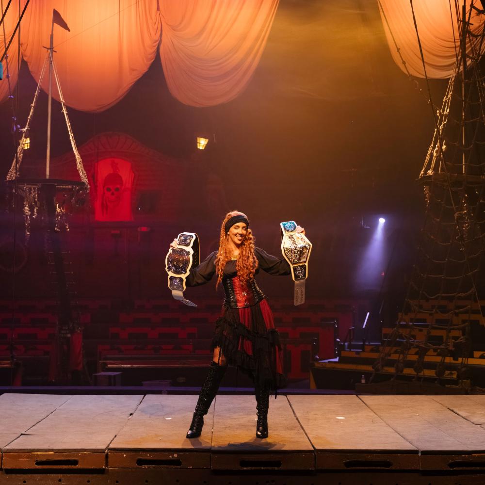 Chelsea Green dressed as a pirate on stage at Pirates Dinner Adventure in Kissimmee, proudly holding two WWE championship belts.