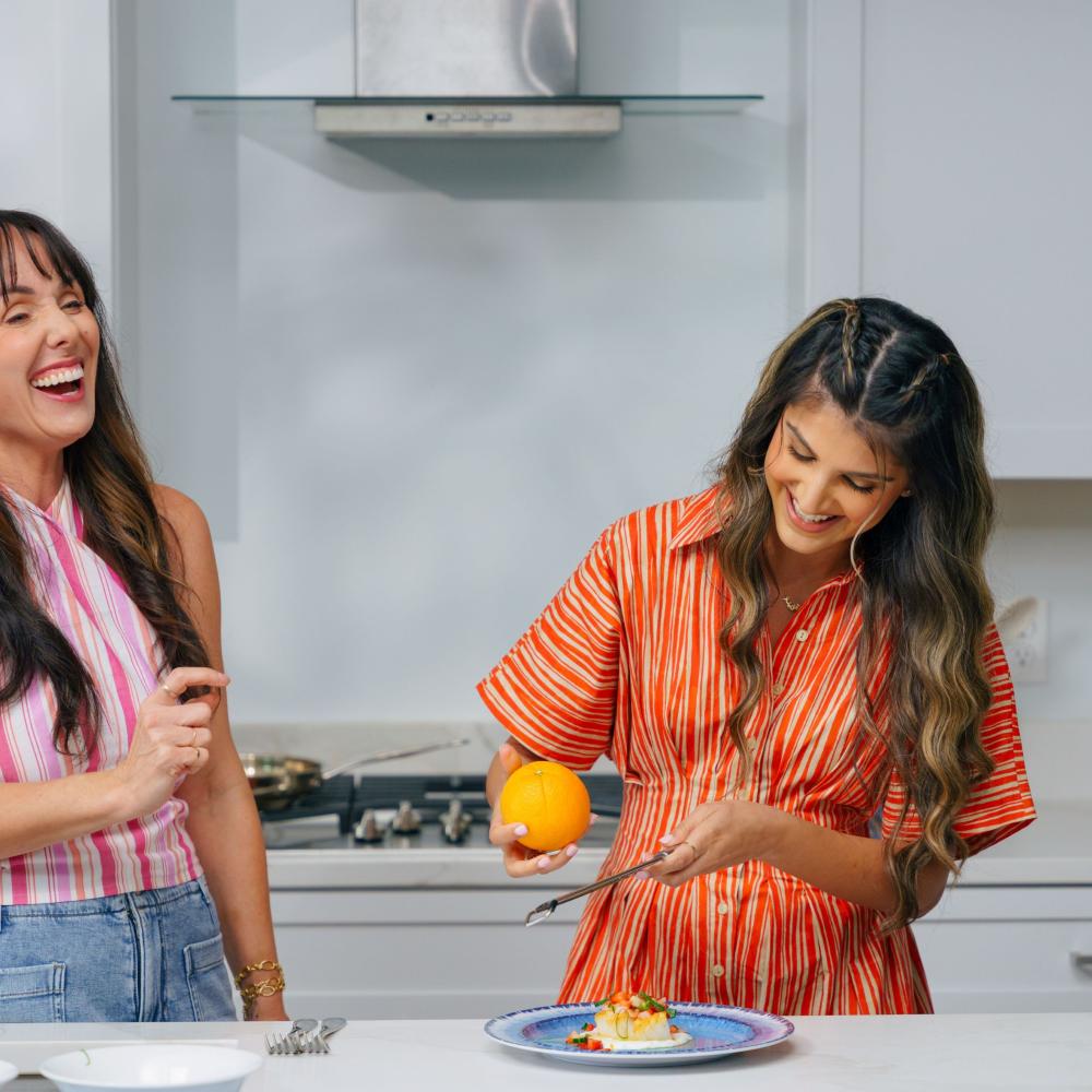 Home chef Maryam Ishtiaq and host Jen Bonner laughing together in a bright, modern Kissimmee vacation home kitchen as they prepare a fresh, colorful dish.